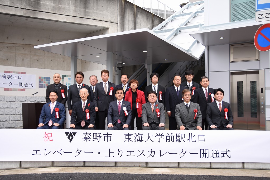 「祝 秦野市東海大学前駅北口エレベーター・上りエスカレーター開通式」と書かれた垂れ幕の前に、スーツを着た複数の関係者が集まって記念撮影をしている写真