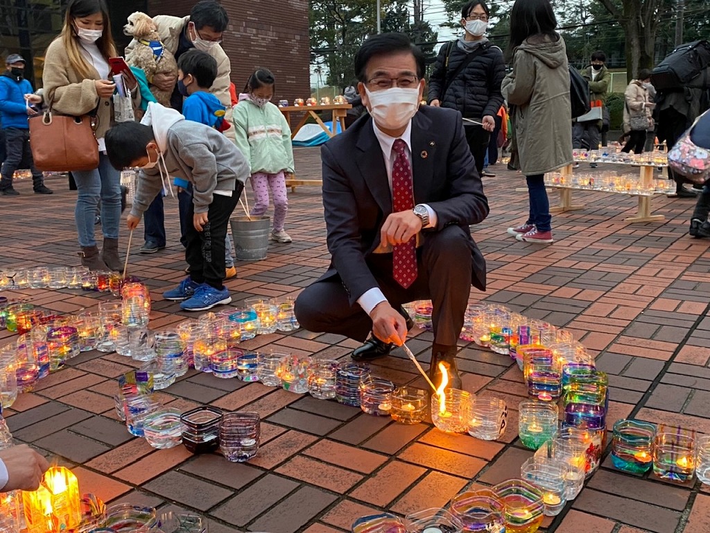 レンガの地面に並べられたグラスの中のキャンドルにかがんで火を灯している、笑顔の高橋市長を正面から撮影した写真