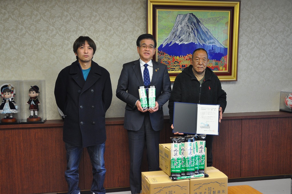中央の高橋市長がパッケージに入った蕎麦を両手に持ち、その両脇に立つ2名の男性とともに記念撮影をしている様子を写した写真