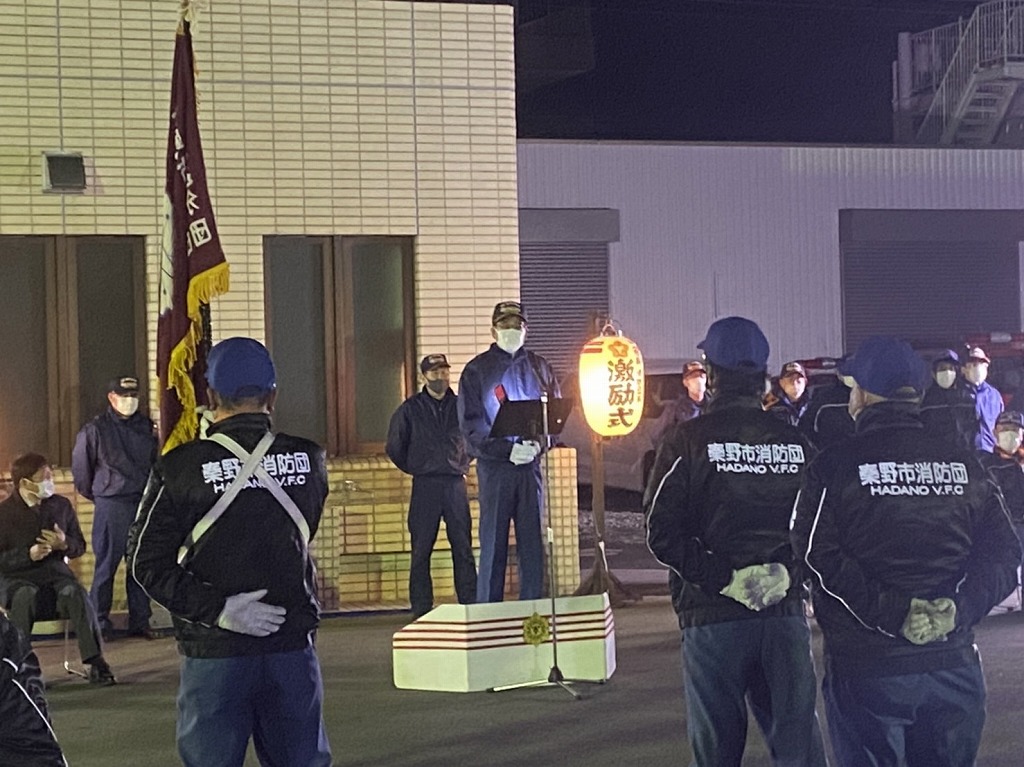 夜の屋外で秦野市消防団のジャンパーを着た複数の男性が整列しており、演台に立つ男性がスピーチをしている消防団歳末火災特別警戒激励式の様子を写した写真