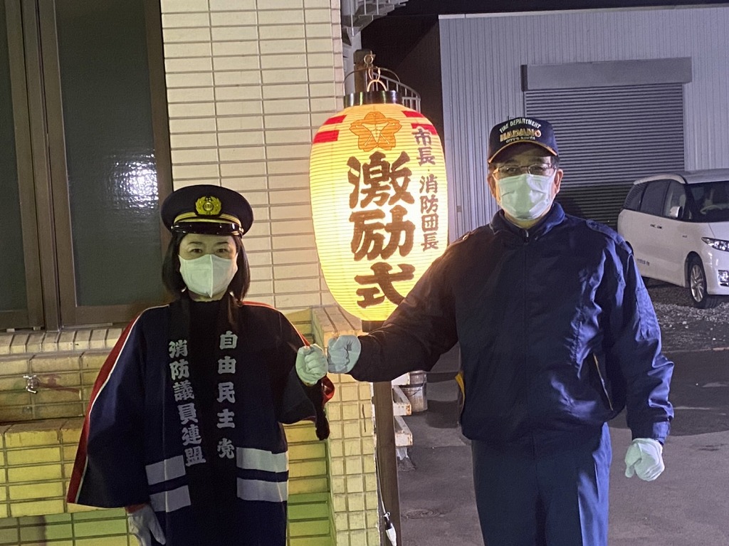 法被を着て帽子を被った女性とキャップを被った高橋市長が、提灯の「市長 消防団長 激励式」と書かれた文字を挟んで拳を合わせ、カメラに視線を送る様子を写した写真