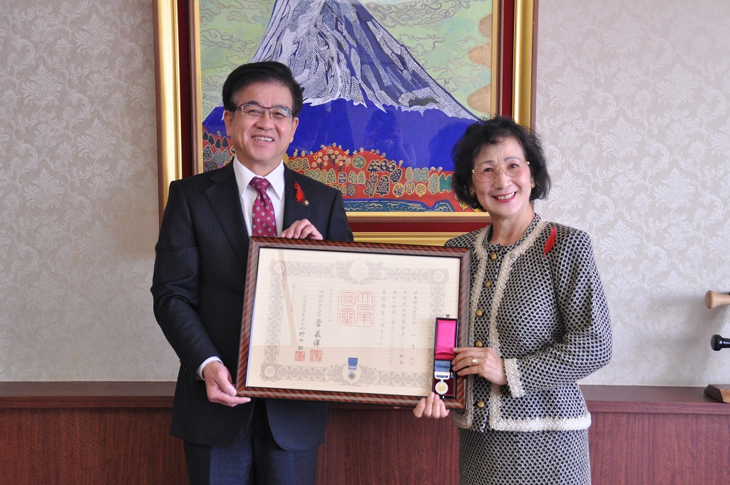 富士山の絵画が飾られた部屋で、スーツ姿の市長と、額縁に入った表彰状と勲章のようなメダルを持った女性が並んで笑顔で写っている写真
