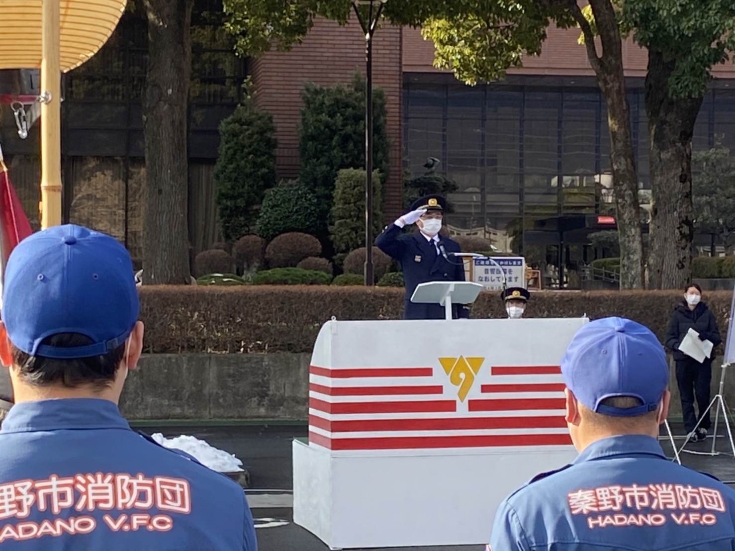 晴れた日の屋外で、秦野市消防団の団員が立っている前で、制服を着た市長が演台の前で敬礼をしている写真
