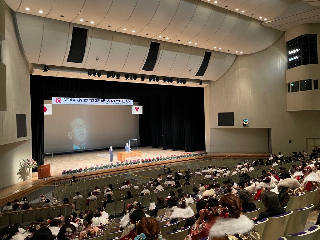 「秦野市新成人を祝うつどい」の横断幕が掲げられた大きなステージが設置されたホールで、振袖や華やかな衣装を着た大勢の新成人が集まり、スクリーンに映し出された映像を見ている写真