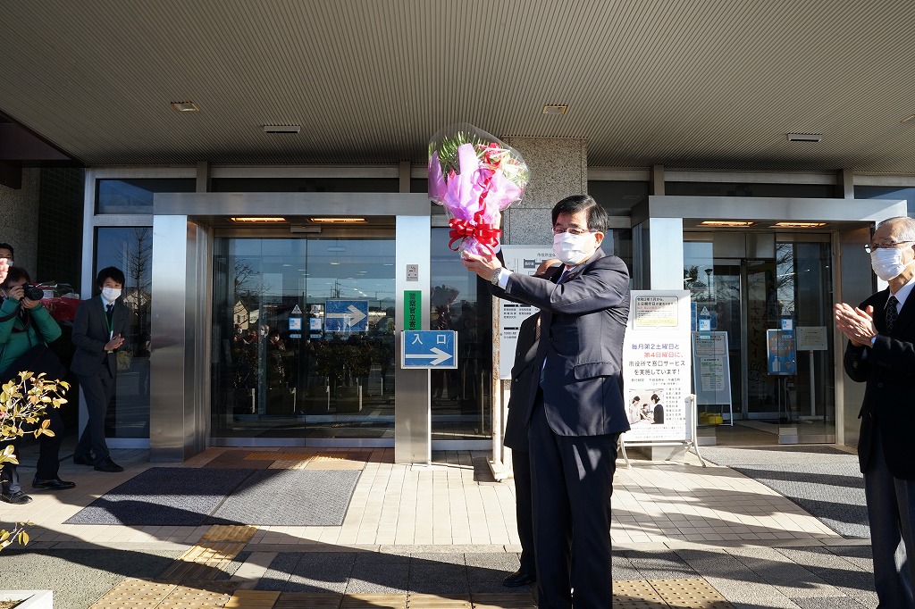 建物のガラス扉の前で、マスクを着用したスーツ姿の市長が、ピンクのラッピングがされた花束を頭上に掲げ、複数の人に囲まれている写真