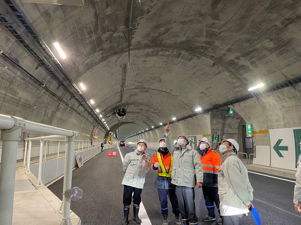 照明で明るく照らされた長いトンネルの中で、ヘルメットを被った複数の作業員が、天井を指差しながら話し合っている工事現場の写真