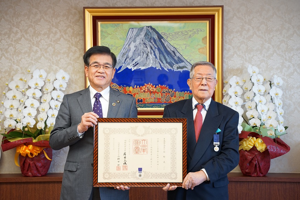 富士山の絵画が飾られ、白い胡蝶蘭が置かれた部屋で、スーツ姿の市長と1人の男性で、額縁に入った表彰状を並んで持ち、笑顔で立っている写真