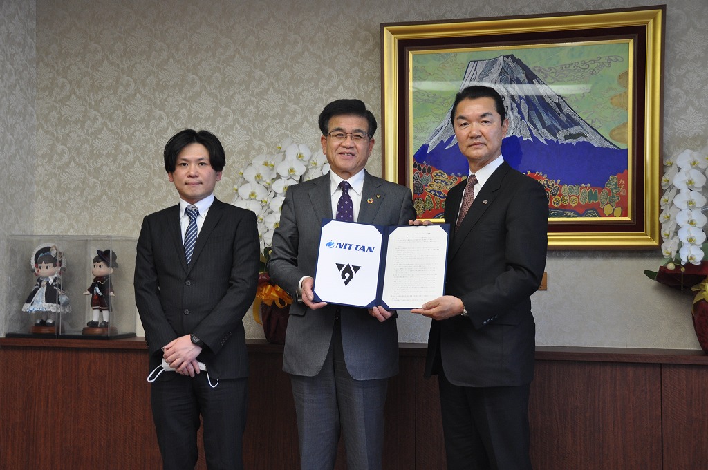 富士山の絵画が飾られ、白い胡蝶蘭が置かれた部屋で、市長を中央にスーツ姿の3人の男性が立ち、市長と1人の男性で会社のロゴ入りの書類と契約書が入ったファイルを持っている写真