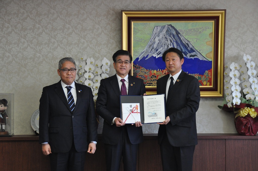 富士山の絵画が飾られ、白い胡蝶蘭が置かれた部屋で、市長を中央にスーツ姿の3人の男性が立ち、市長と1人の男性で目録と感謝状が入ったファイルを持っている写真
