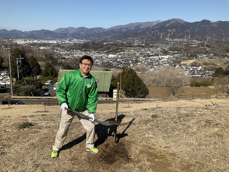 奥に街並みと山々が広がる風景が見える晴れた冬の丘の上で、緑のジャンパーを着た市長がスコップを持ち、植樹活動をしている写真
