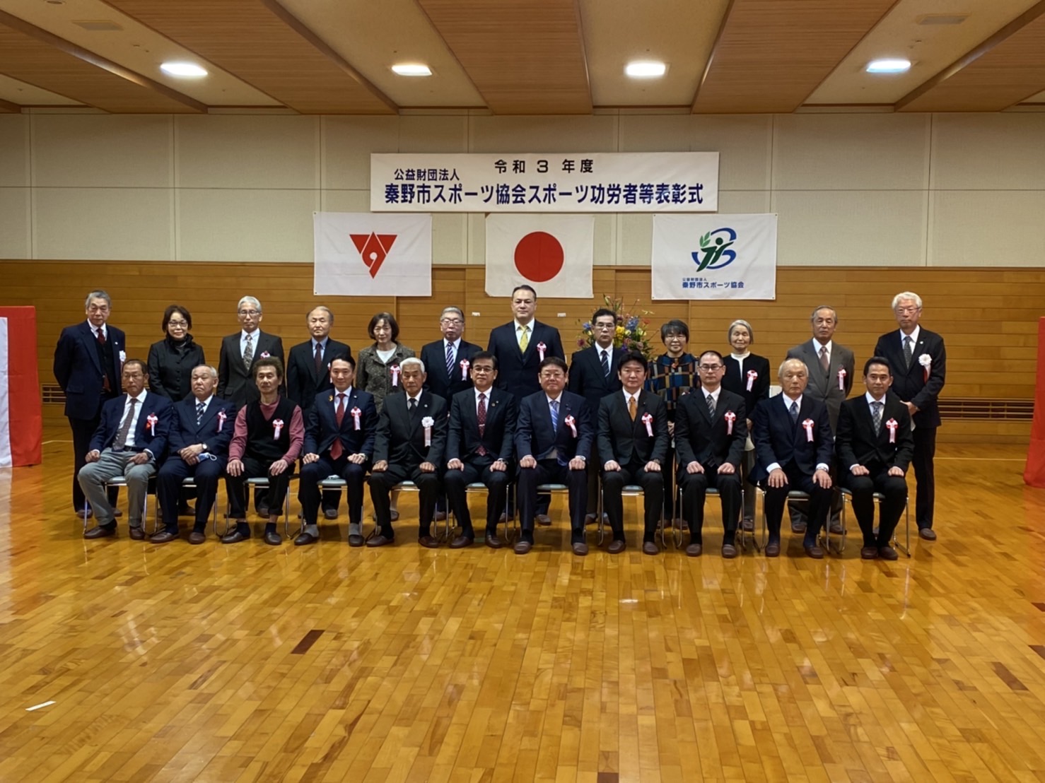 「公益財団法人秦野市スポーツ協会スポーツ功労者等表彰式」の横断幕と国旗・市章旗の前で、胸章をつけた男女が並んで写っている表彰式の集合写真