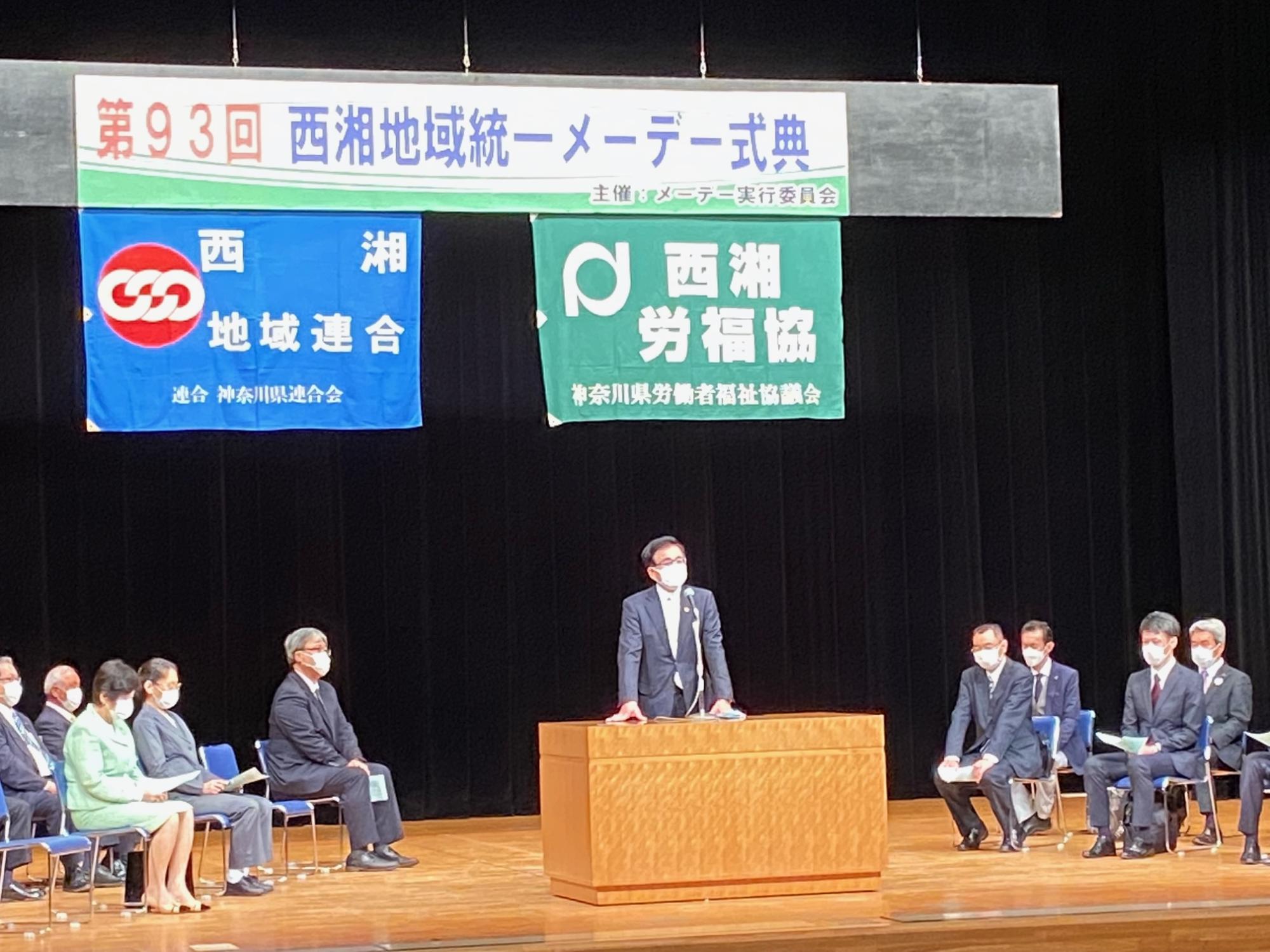 多くの人々が座って見守る中、壇上の木製演台に立つ市長がマイクを前にスピーチしている様子の写真