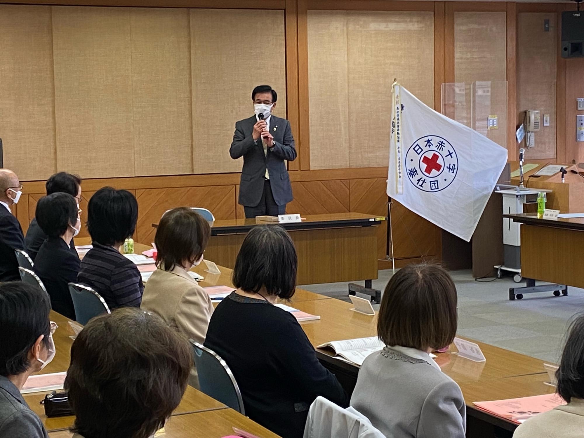 木製のテーブルと椅子が並ぶ会議室で、日本赤十字社の旗が立つ横で市長が聴衆に向けてマイクで話している様子の写真