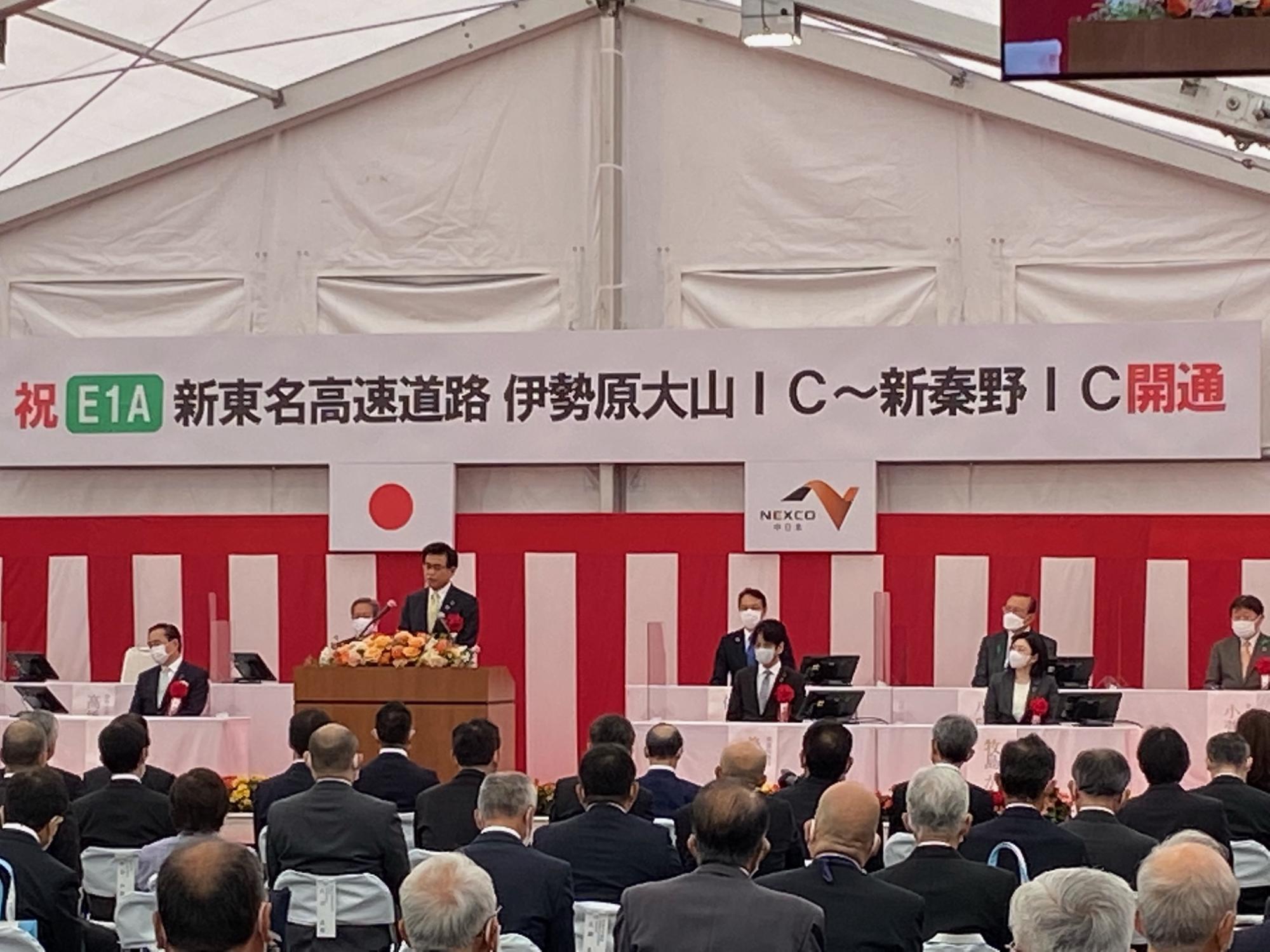 後方に新東名高速道路の開通を祝うメッセージが書かれている横断幕と紅白幕の装飾がされたテント会場で、壇上の市長がマイクを持ちスピーチしている写真