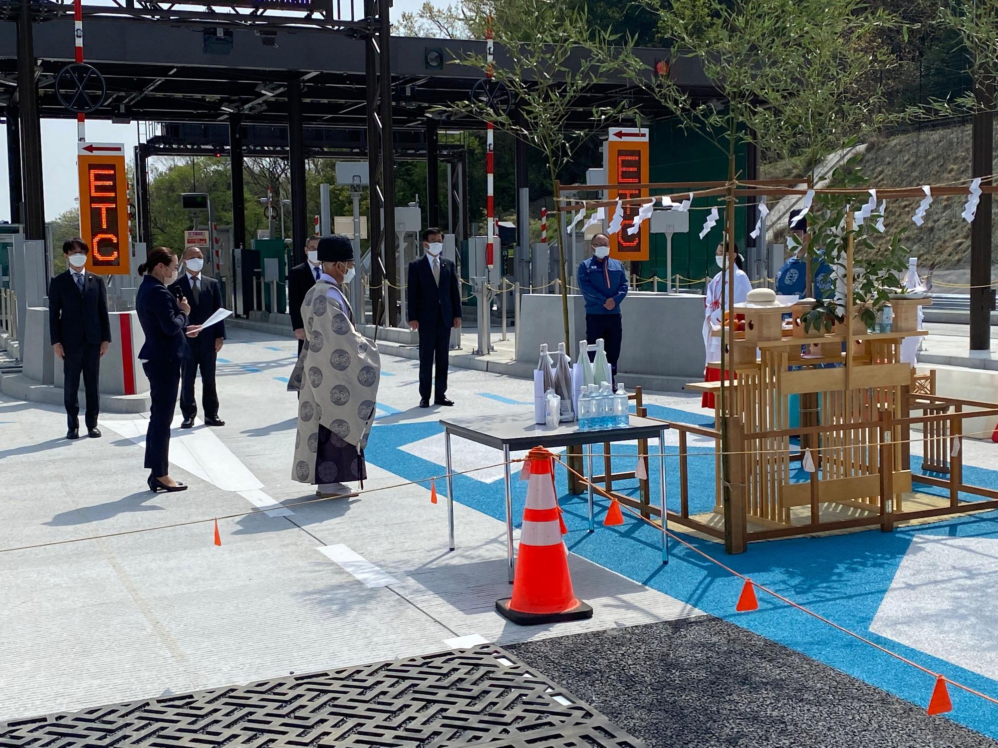 新しい料金所の前に供え物が並ぶ祭壇が設けられ、関係者や神主が参列し、神事が行われている様子を捉えた写真