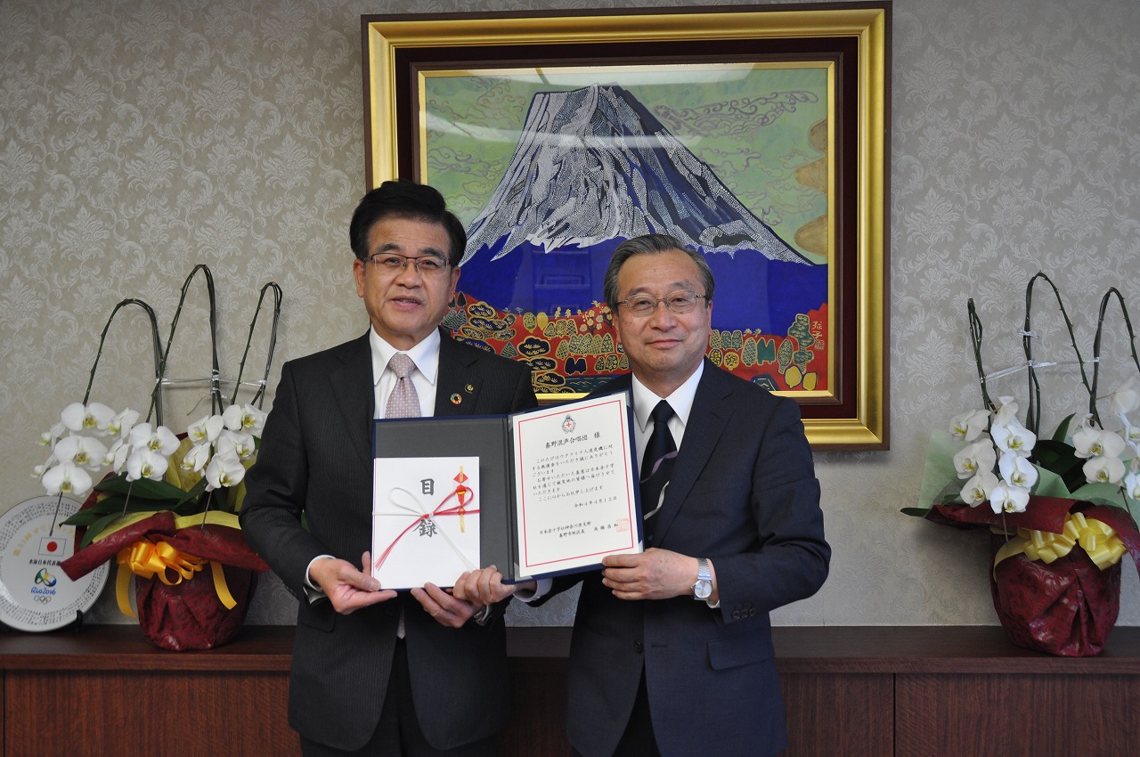 富士山の絵画が飾られた室内で、スーツ姿の市長と男性が寄付目録と感謝状を掲げて、笑顔でカメラに向かって立っている写真