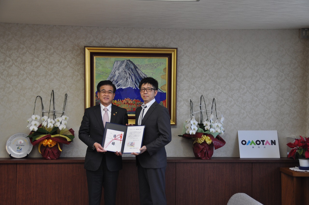 富士山の絵画が飾られた室内で、スーツ姿の市長と男性が寄付目録と感謝状を掲げ、カメラに向かって立っている様子の写真