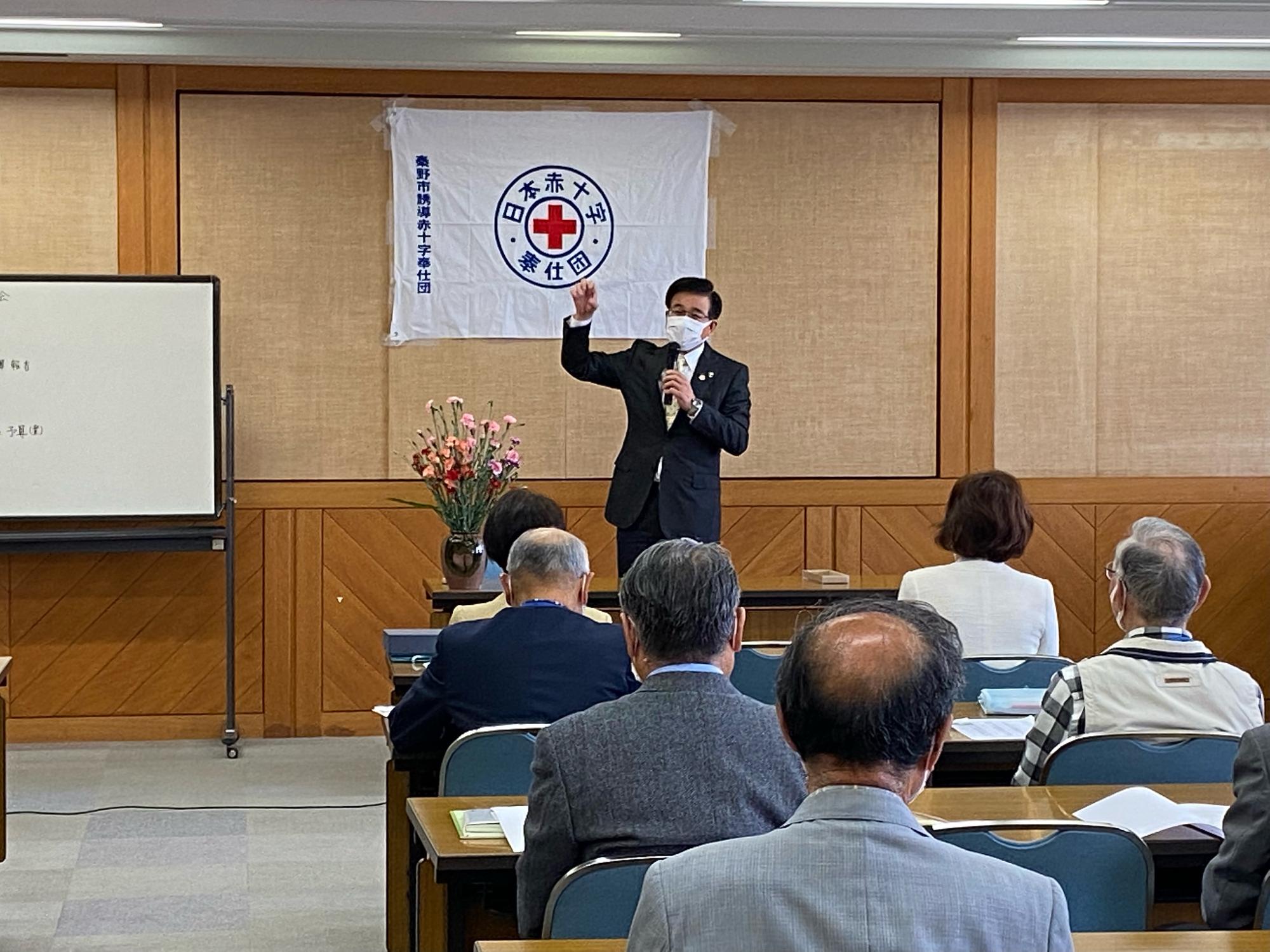 白地に日本赤十字社のロゴが描かれた旗が掲げられた部屋で、スーツ姿の市長がマイクを持って聴衆に身振り手振りを交えながら話している会議の様子の写真