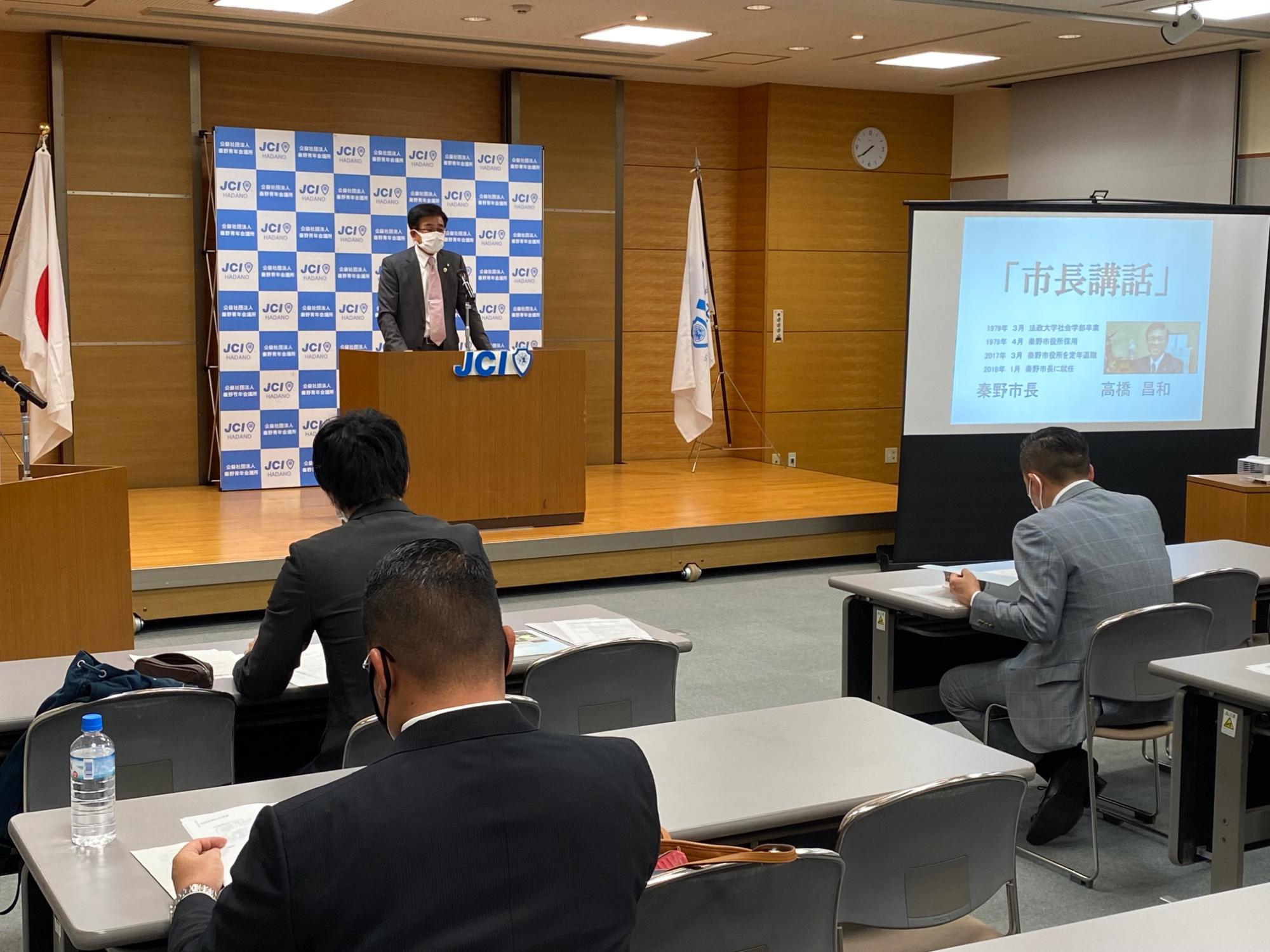 背後にJCIのロゴが入ったバックパネルや横に「市長講話」と映し出されたスクリーンがあり、木製の演台に立つスーツ姿の市長が、多数の聴衆の前で話している会議の様子の写真