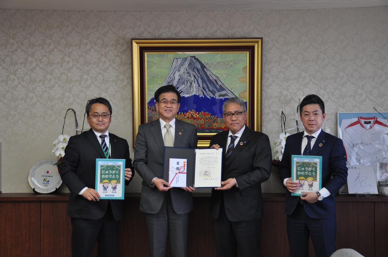 富士山の絵画が飾られた室内で、スーツ姿の市長と3人の男性が並んで、市長と男性で感謝状と寄付目録、両端の男性は「にゅうがくおめでとう」と書かれたファイルを掲げている写真
