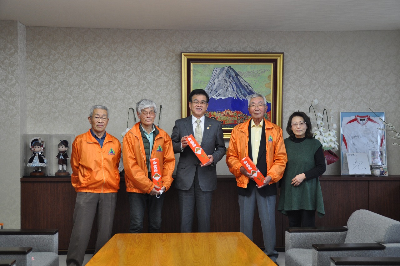 富士山の絵画が飾られた室内で、中央のスーツ姿の市長を囲んで、オレンジ色の上着を着た3人の男性と1人の女性が並び、そのうち市長と男性2人が赤い箱を手にしている写真