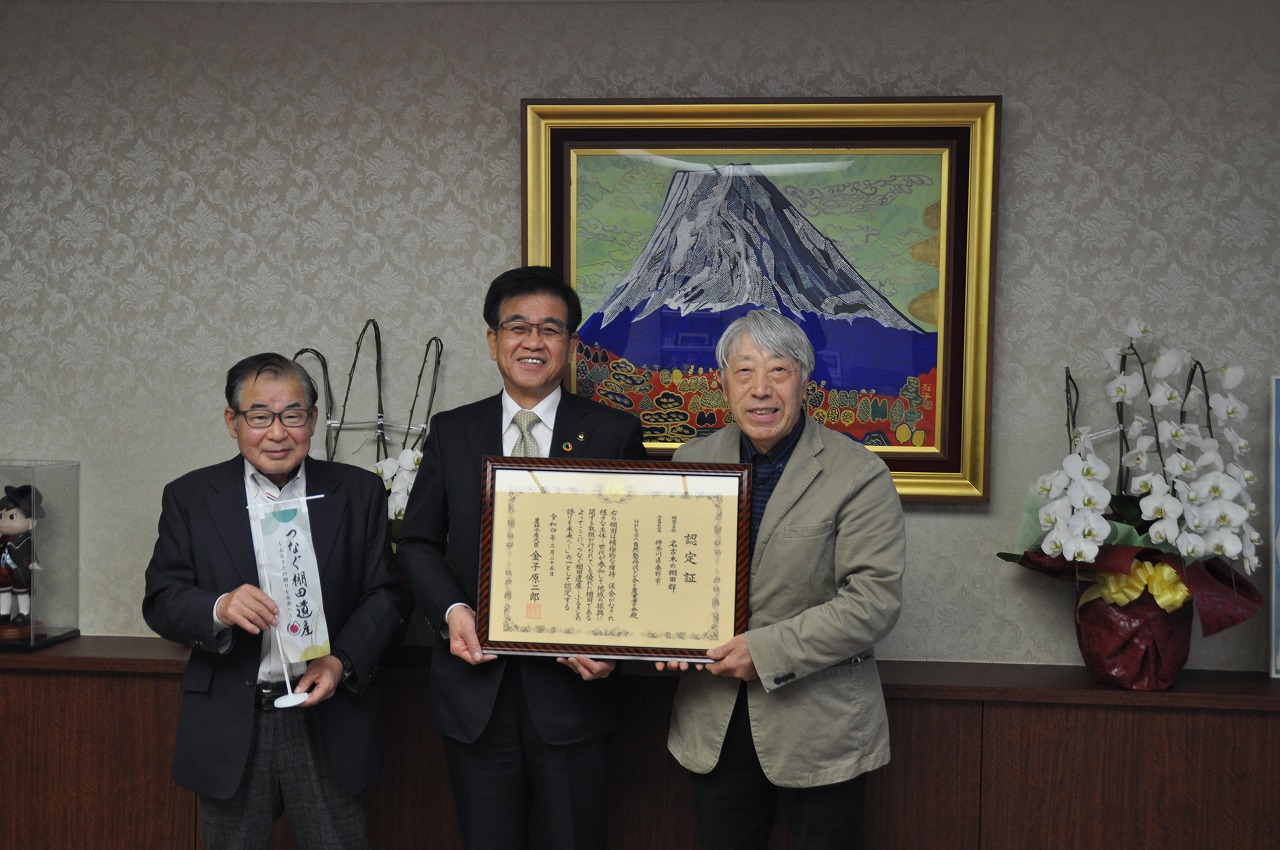 富士山の絵画が飾られた室内で、スーツ姿の市長と男性で認定書を掲げ、隣の男性が小さい旗を持って並んで立っている写真