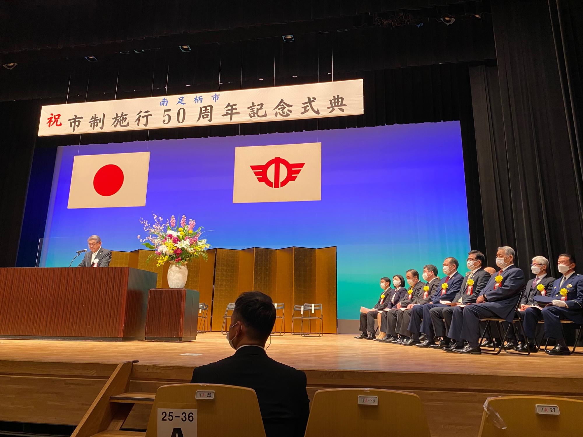 「祝 市制施行50周年記念式典」の横断幕の下で、日本の国旗と市章が掲げられた壇上で男性がスピーチしており、多くの来賓が並んで着席している写真