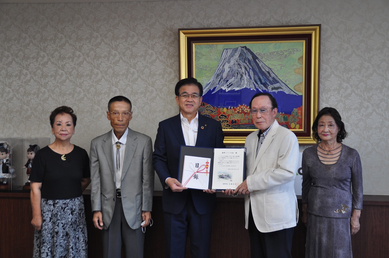 富士山の絵画が飾られた部屋で、市長の両脇に男女2人ずつが立ち、市長と男性で目録と感謝状が入ったファイルを持って立っている写真