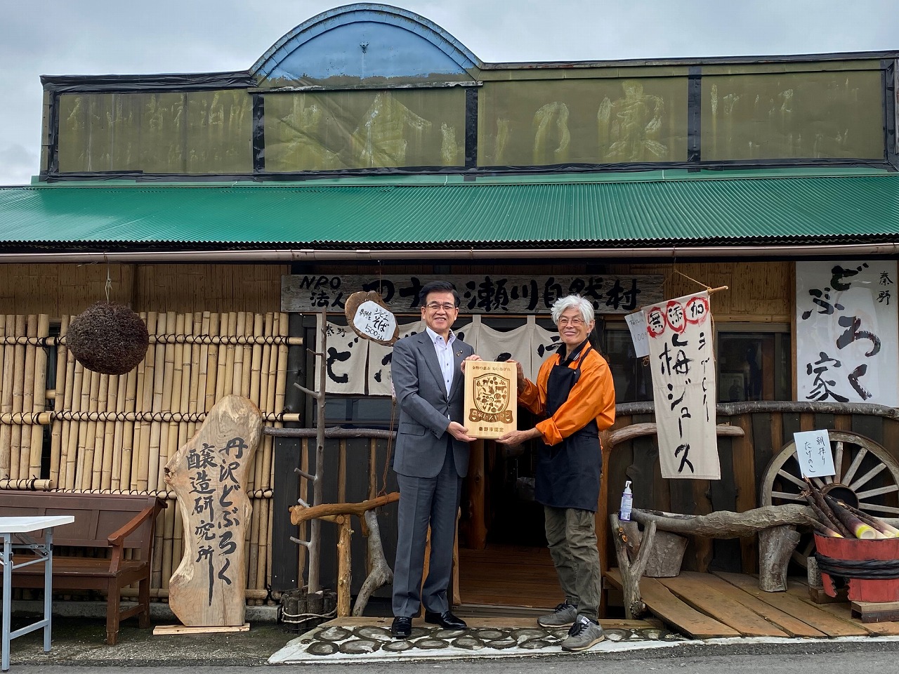 古民家風の建物の前で、スーツ姿の市長とオレンジ色の服を着用した男性が、木製の看板を並んで持っている写真