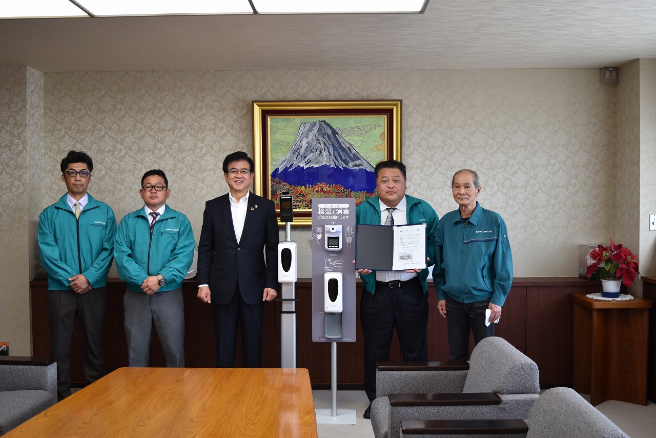 富士山の絵画が飾られた部屋で、スーツ姿の市長が、職員らしき制服姿の4人の男性に囲まれ、検温・消毒器と感謝状を挟んで並んでいる贈呈式の写真