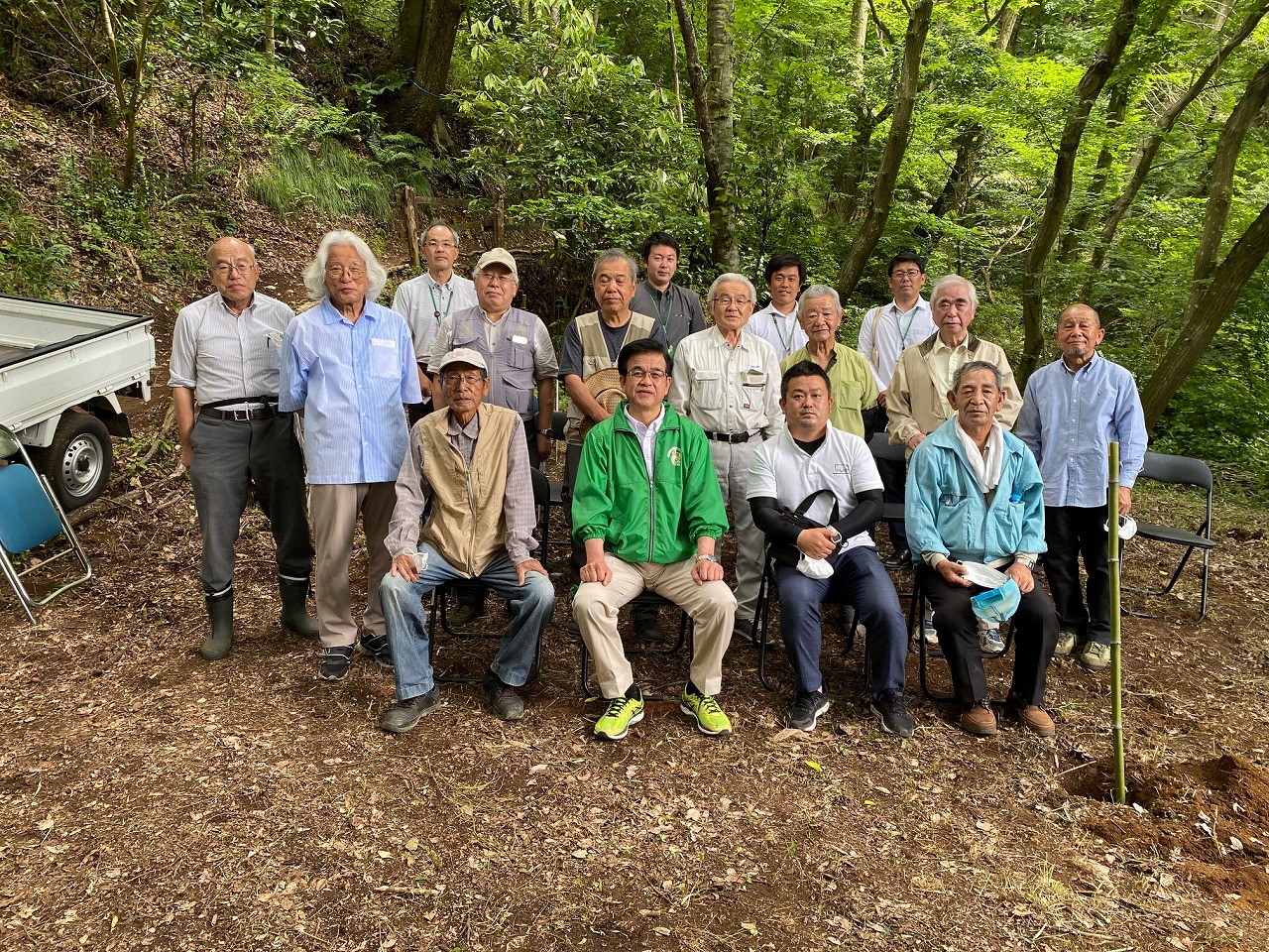 森の木々を背景にした広場で、緑色のジャンパーを着た市長を中央に植樹に参加した多数の男性が集合して写した写真