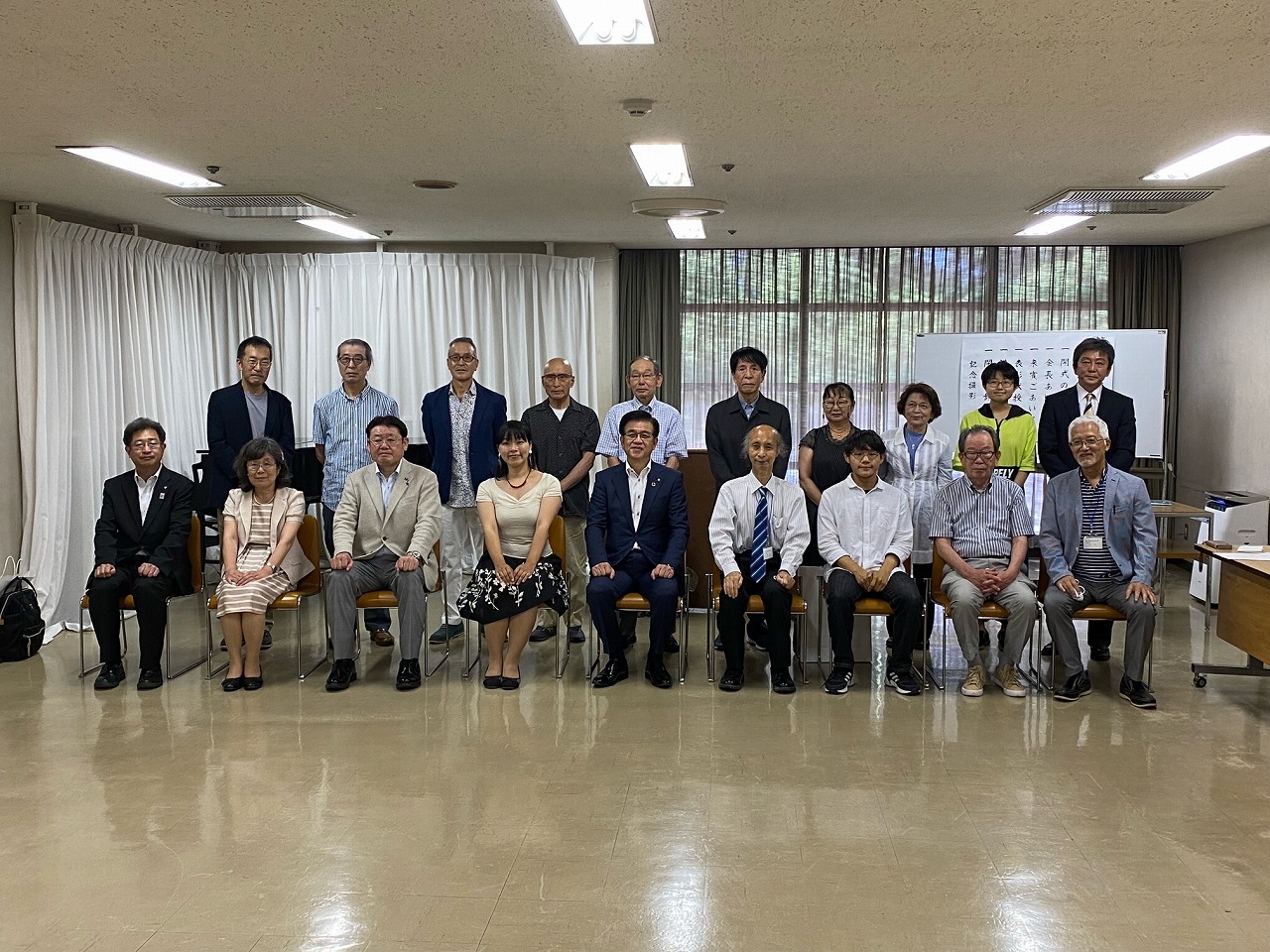 会議室のような部屋で、市長を中央にしてスーツ姿やカジュアルな服装の男女が、座っている人や立っている人2列に分かれて並んでいる集合写真