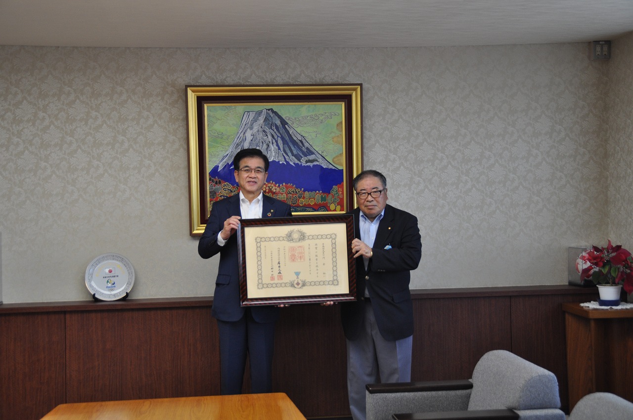 富士山の絵画が飾られた部屋で、市長と男性が、中央で大きな額縁に入った表彰状を並んで持って笑顔を見せている写真