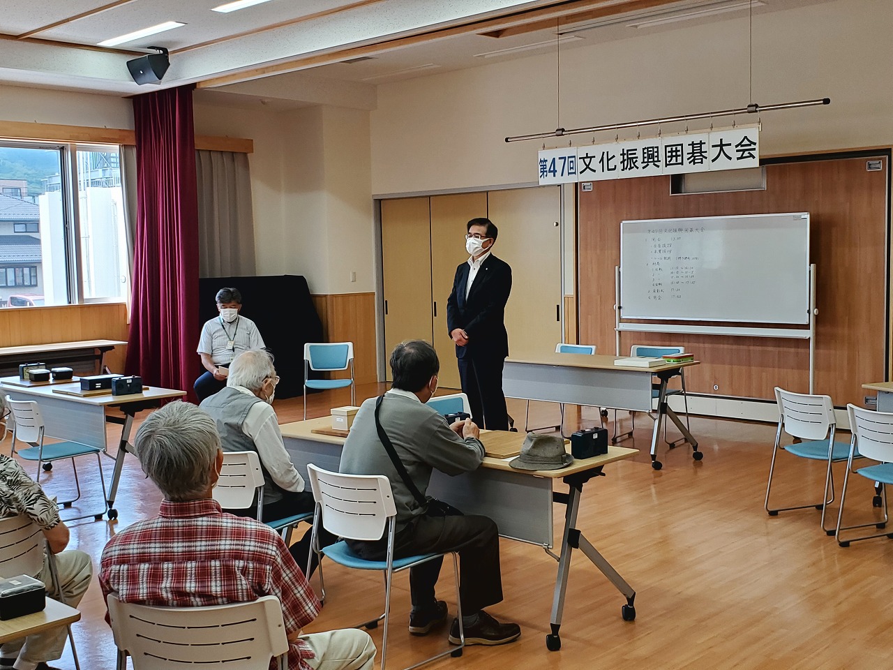 第47回文化振興囲碁大会の会場で、スーツ姿の市長が、参加している複数の高齢者に向かって挨拶をしている写真