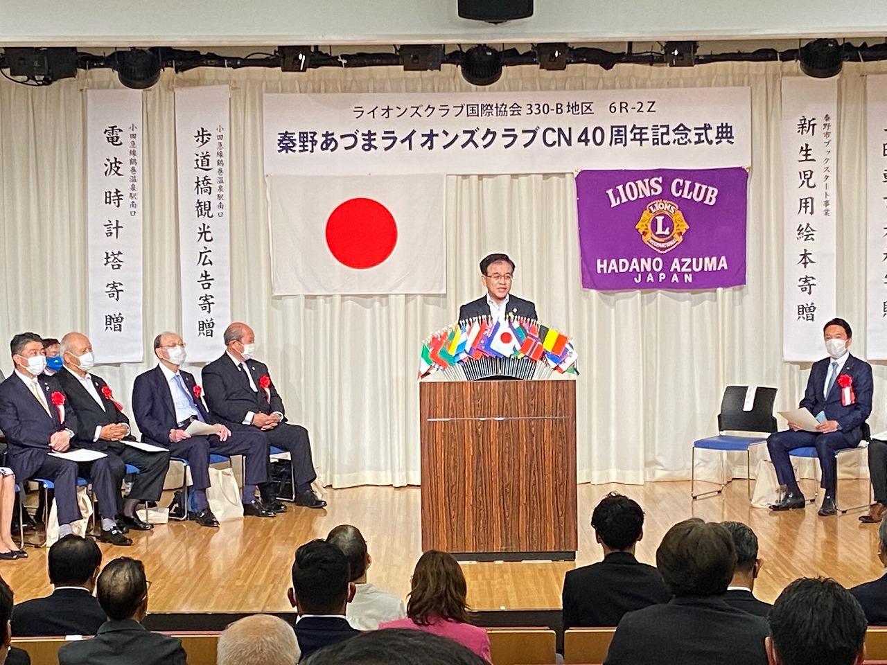 国旗とライオンズクラブの旗が飾られたステージ上で、スーツ姿の市長が、各国旗が飾られた演題でたくさんの観客へ向けてスピーチをしている記念式典の写真