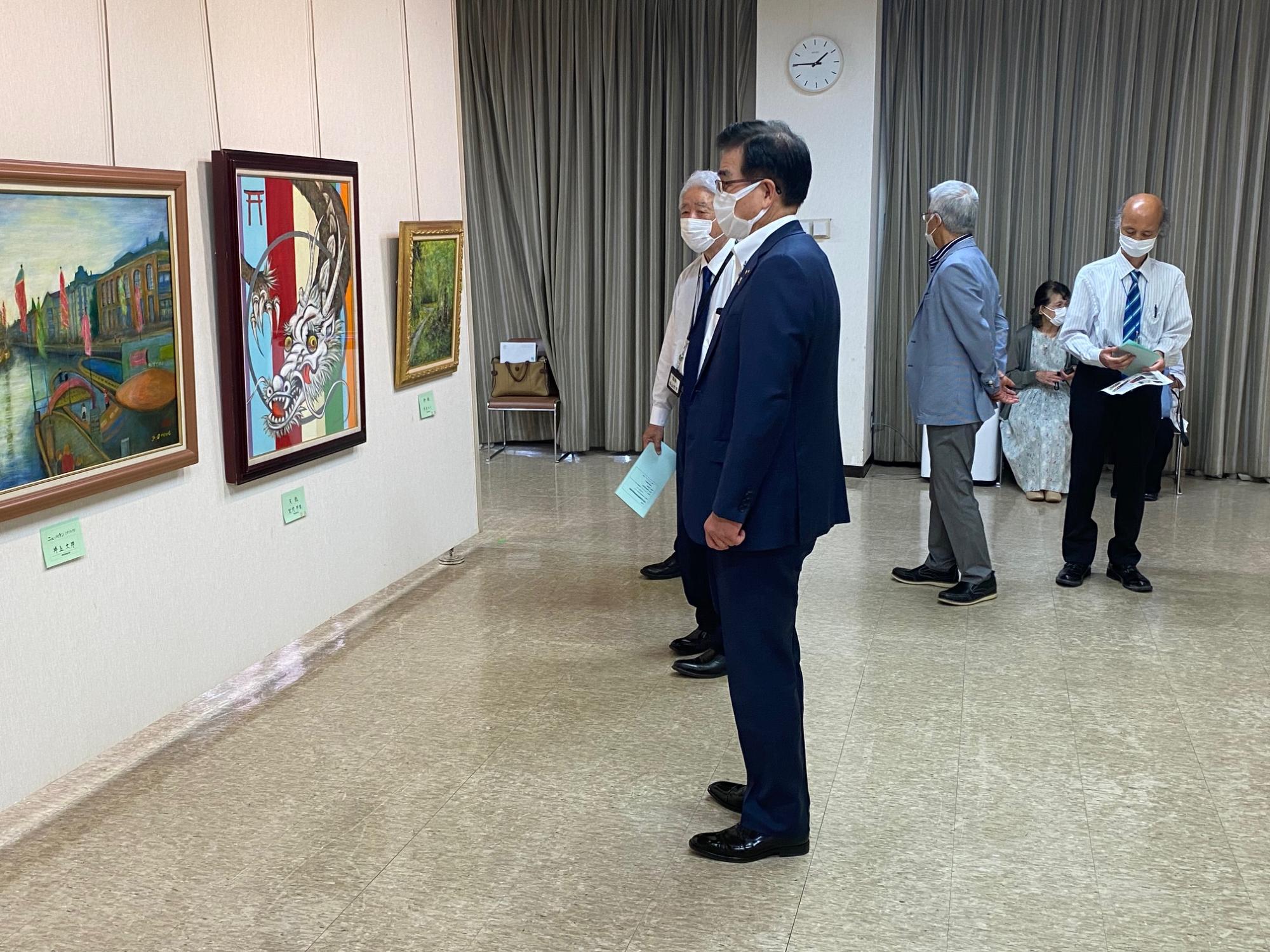 スーツ姿の市長が、風景画や龍の絵が展示された壁の前で、他の来場者や係員と共に鑑賞している美術展の写真