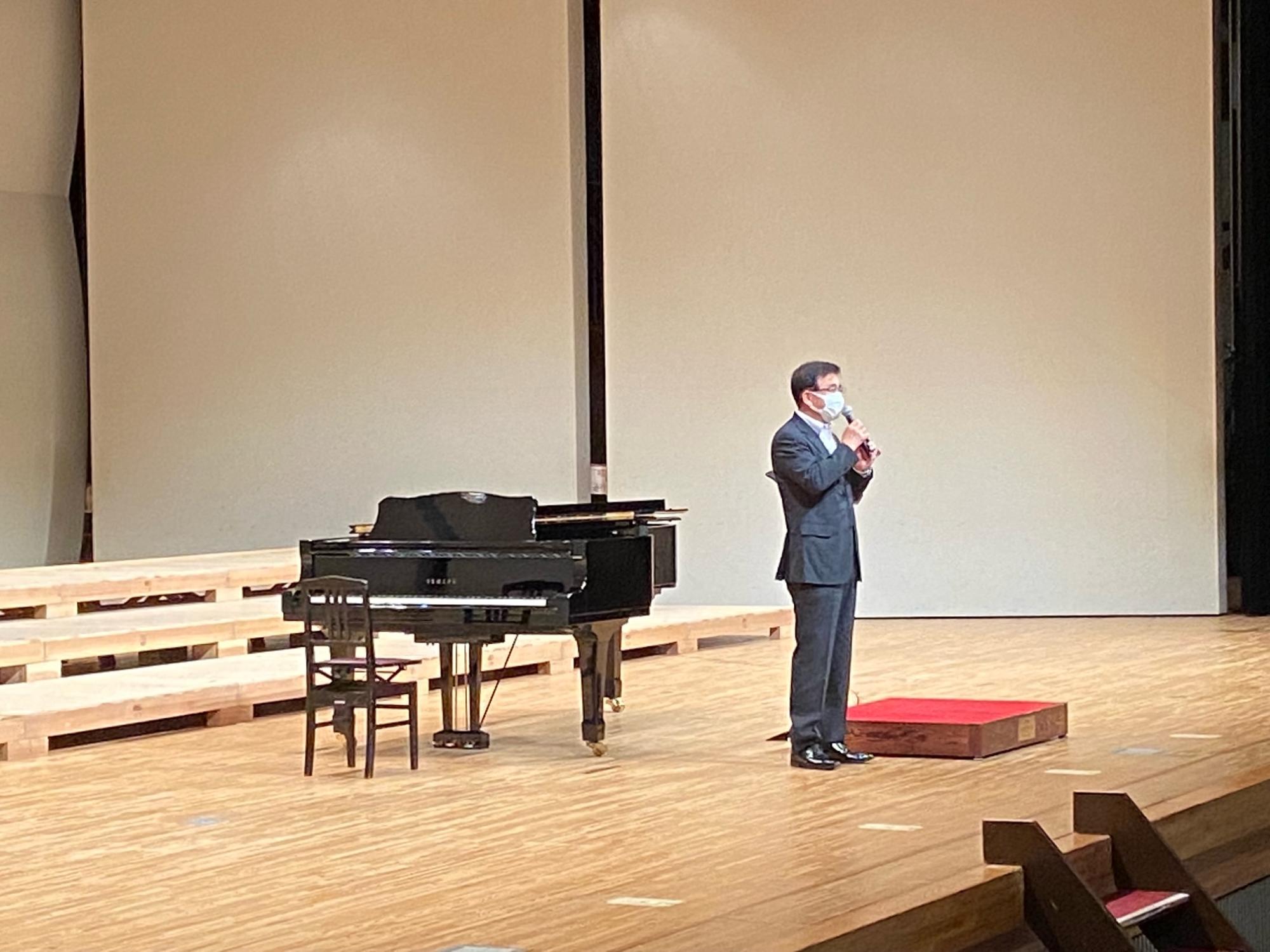 グランドピアノが置かれているコンサートホールのようなステージ上で、スーツ姿の高橋市長がマイクを手にスピーチをしている写真