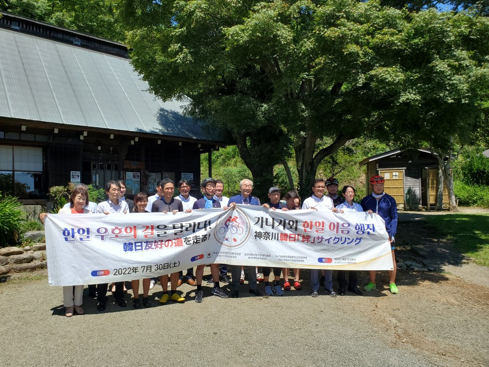 緑の葉を生い茂らせた木の前で、「2022年7月30日」の日付と、韓国語と日本語が記された白い横断幕を男女合わせて十数人が笑顔で持ち記念撮影をしている写真