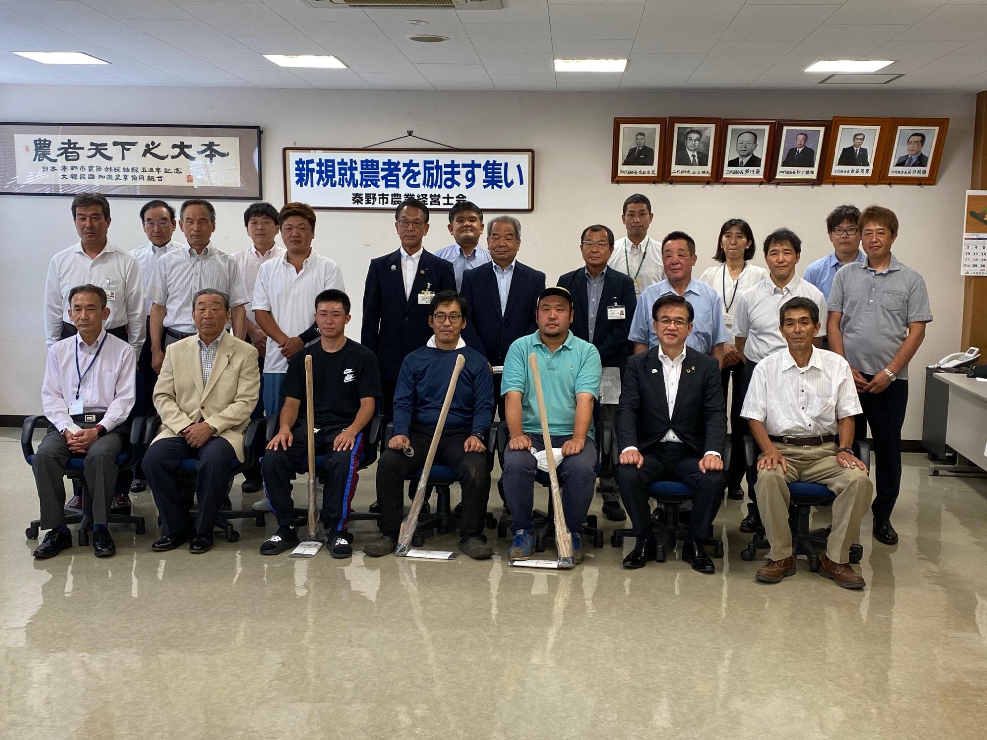 背後に歴代会長の写真、「農者天下之大本」と「新規就農者を励ます集い」と書かれた額が掲げられている室内に大勢の関係者の方が集まり、記念撮影をしている写真