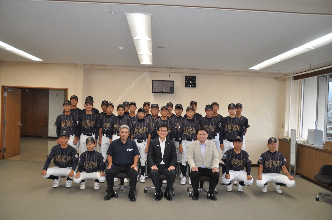 椅子に座っている高橋市長を中心に、両側に関係者の男性2名が座り、周りにユニフォームを着た秦野リトルシニアの選手が集まり集合写真を撮っている写真