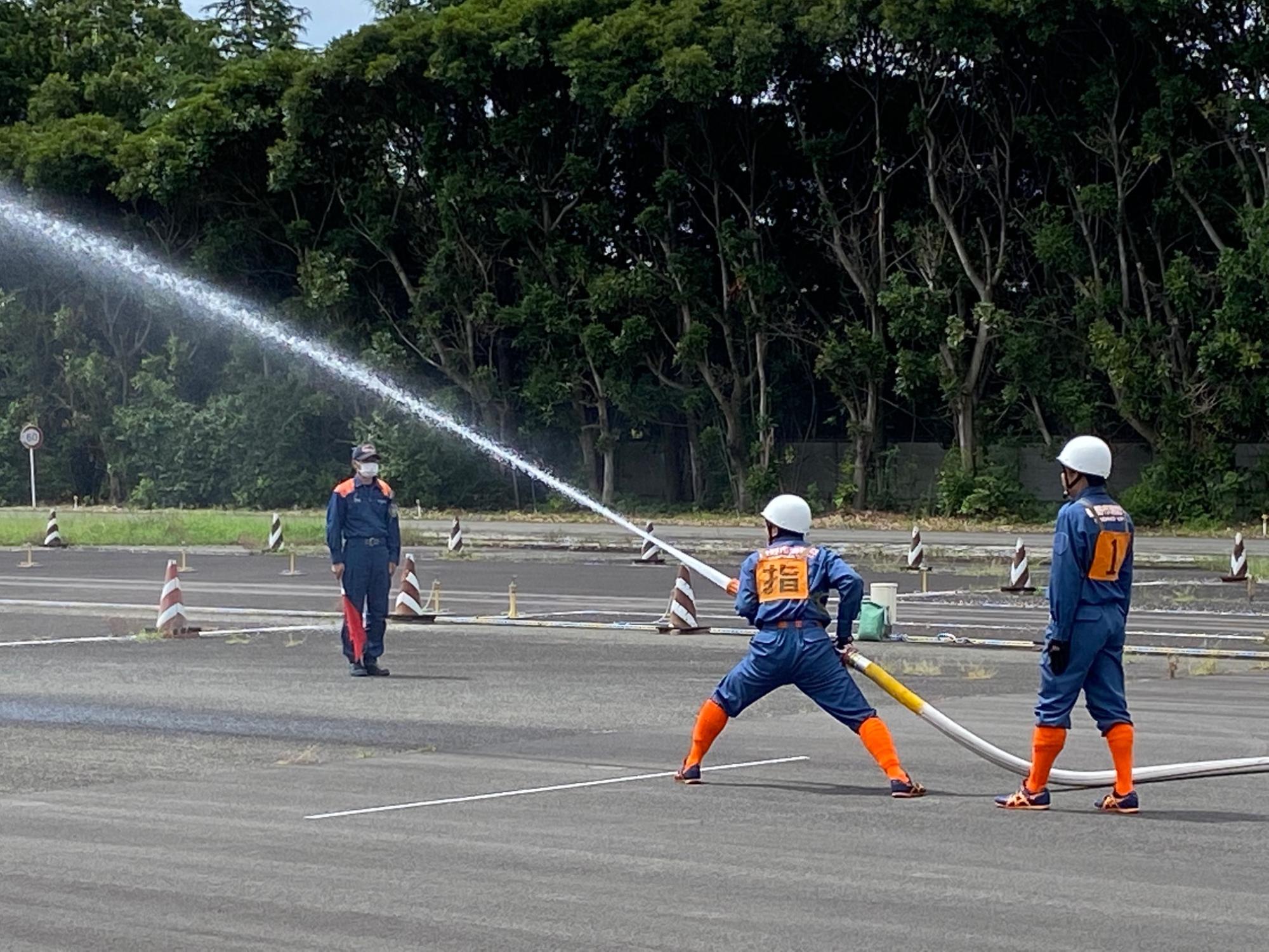 消防服を着た男性がホースから勢いよく水を放水し、離れた場所から赤い旗を持った男性が一人立っている様子を捉えた神奈川県消防操法大会の写真