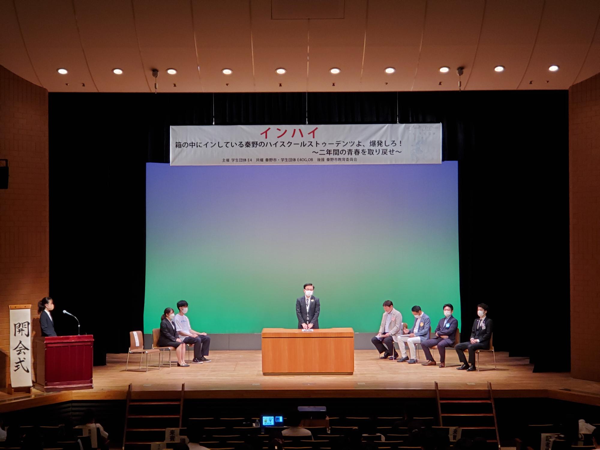 「箱の中にインしている秦野のハイスクールストゥーデンツよ、爆発しろ！二年間の青春をを取り戻せ」と書かれた横断幕の下、演台に立った高橋市長が話をしている開会式の様子の写真