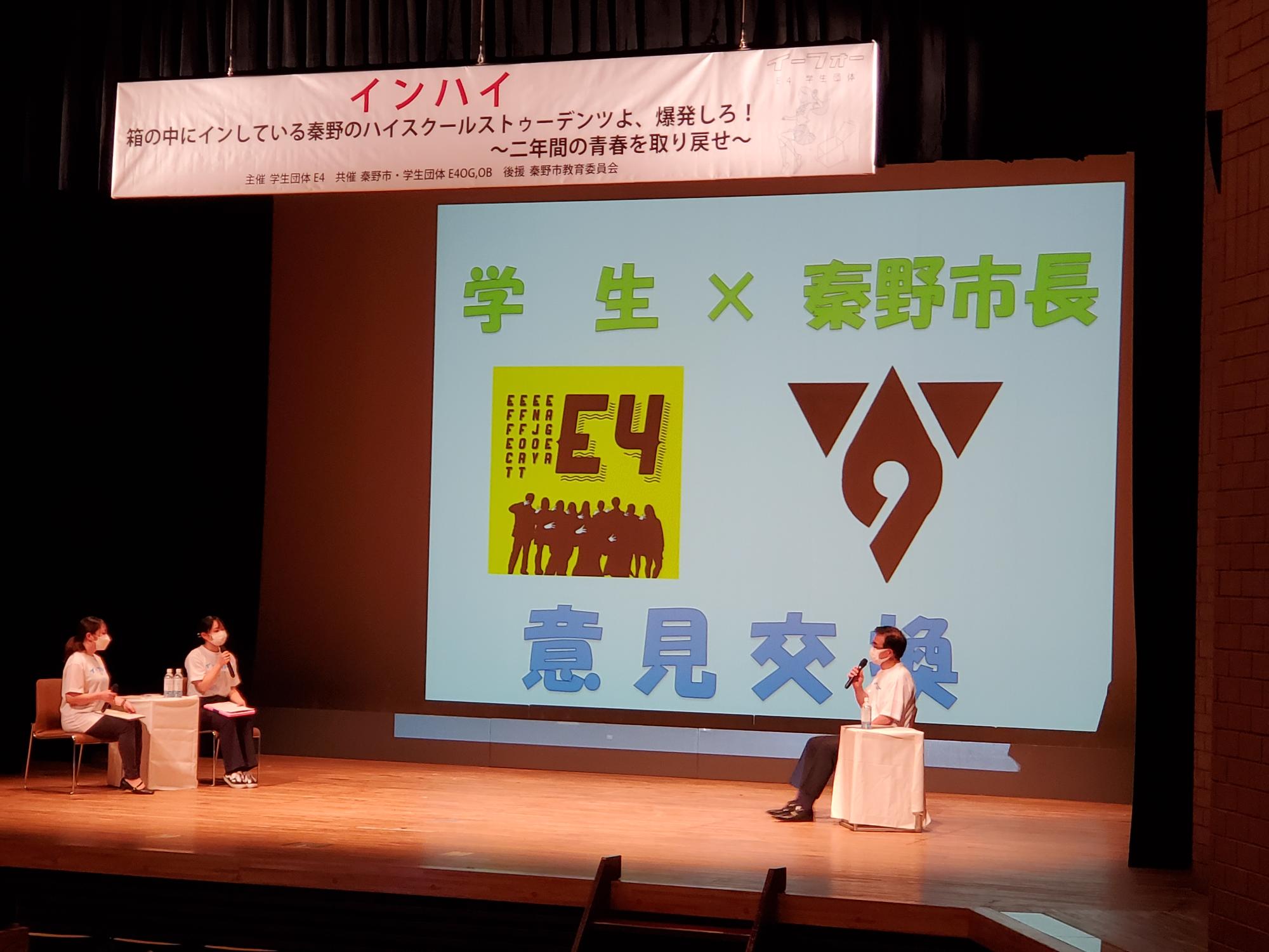 スクリーンに「学生&times;秦野市長 意見交換」と表示され、ステージ右側でマイクを持った高橋市長が椅子に座って話しており、その対面には二人組の女性が座っている意見交換の様子の写真