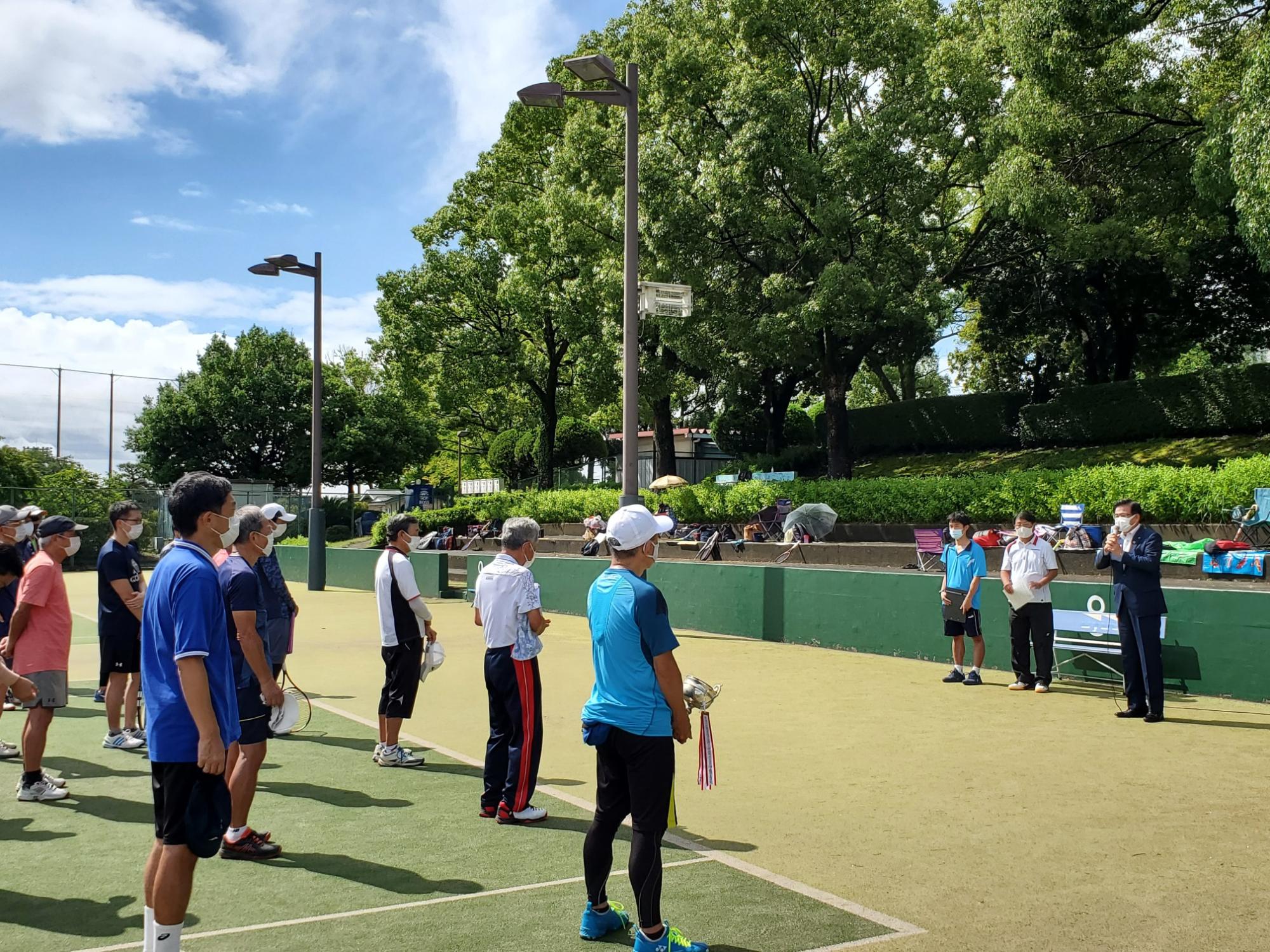 晴天の下、テニスコート上で参加者の方たちが整列し、その先でマイクを持った高橋市長が話をしている様子の写真