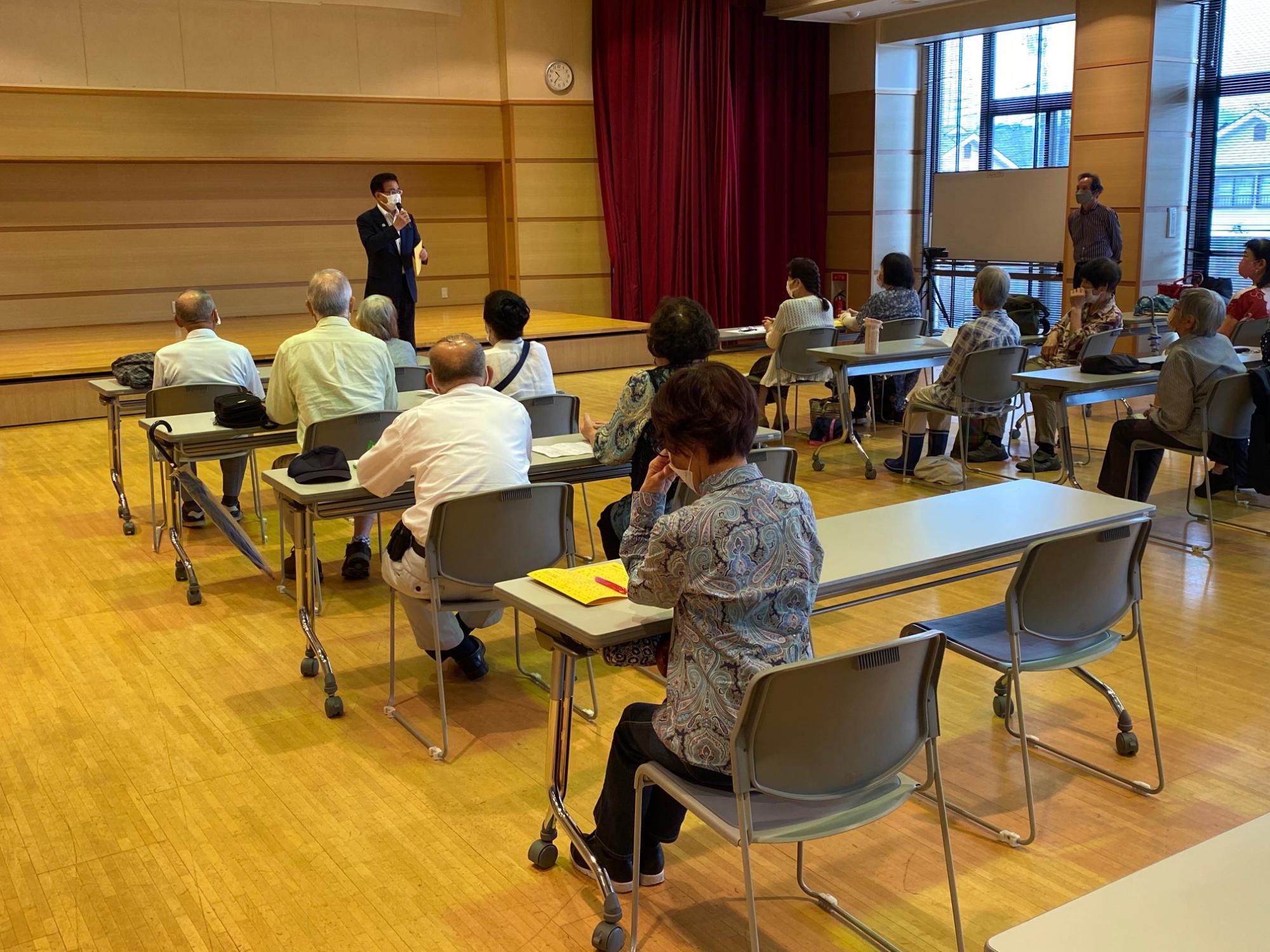 2人掛けの長机が設置された席に参加者たちが座り、前方に立っている高橋市長の話を聞いている様子の写真