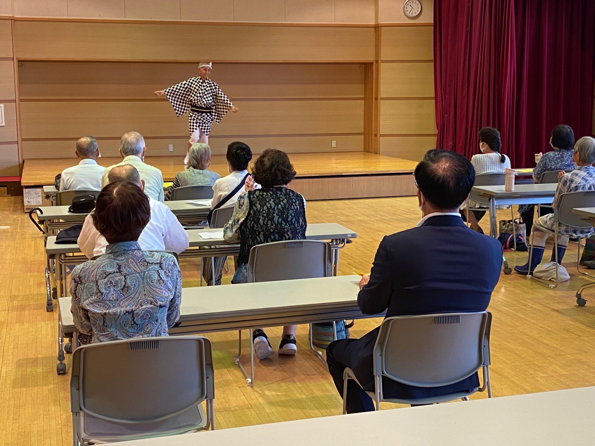 広い室内の小上がりになっている場所で、チェック柄の衣装を身につけた演者が、椅子に座る参加者たちに向かって踊りを見せている様子の写真
