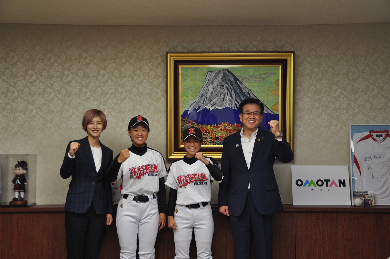 富士山の絵画が飾られた室内で、スーツ姿の市長と女性の間に2人のユニフォーム姿の女子野球選手が立ち、笑顔で拳を突き上げている写真