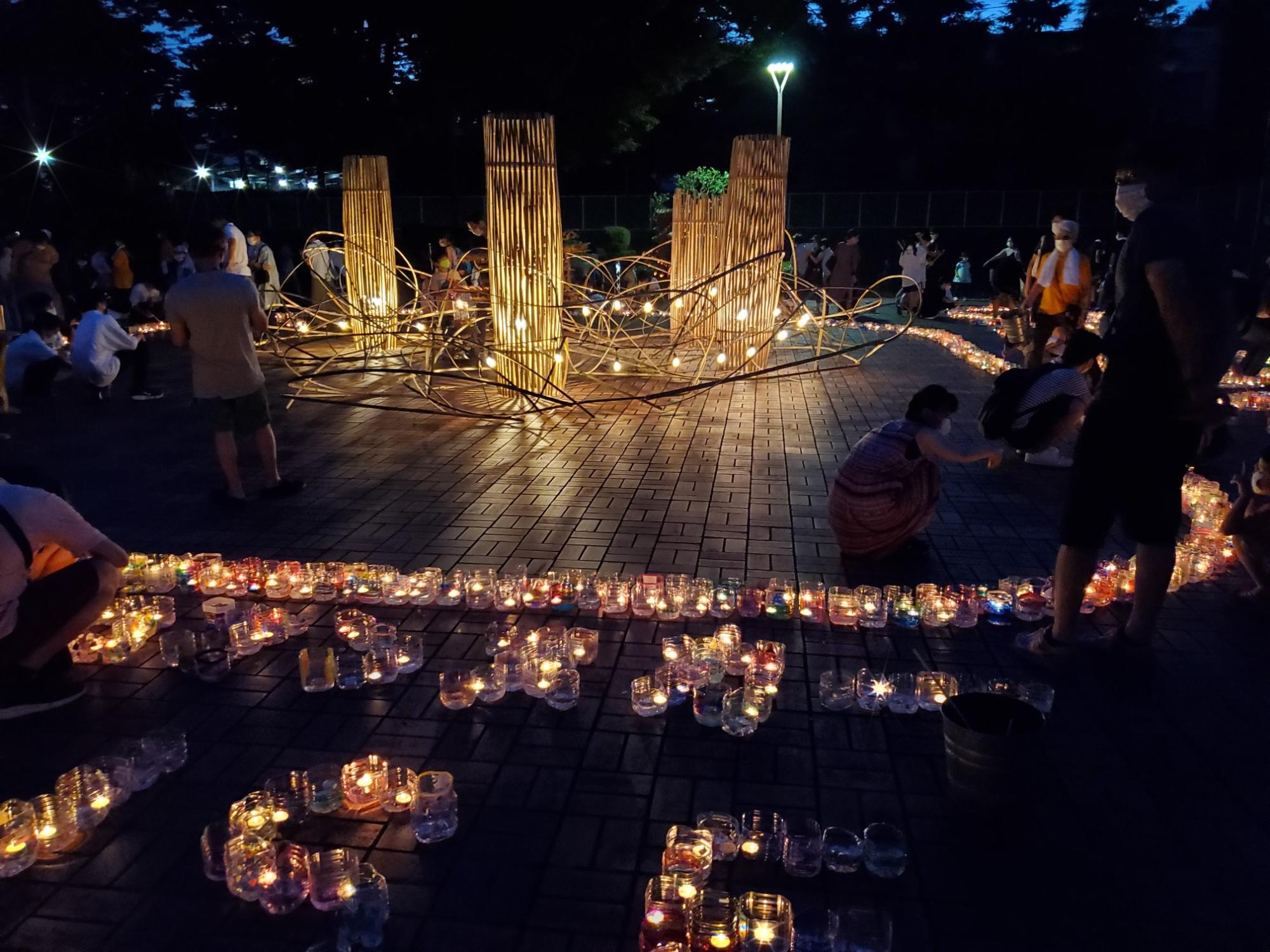 多くの人々が集う暗い公園の広場で、いくつもの灯籠が地面に並べられており、その明かりが幻想的な雰囲気を醸し出している夜祭の写真