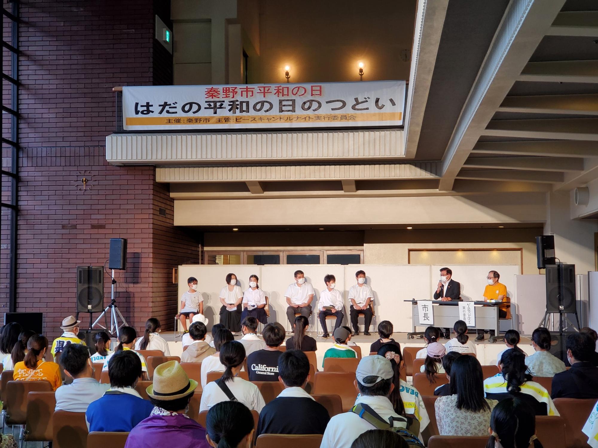 屋内講堂の舞台上で市長がマイクを持ち、横断幕の下に並んだ複数の人々が椅子に着席し、舞台下には多くの聴衆が座っている写真