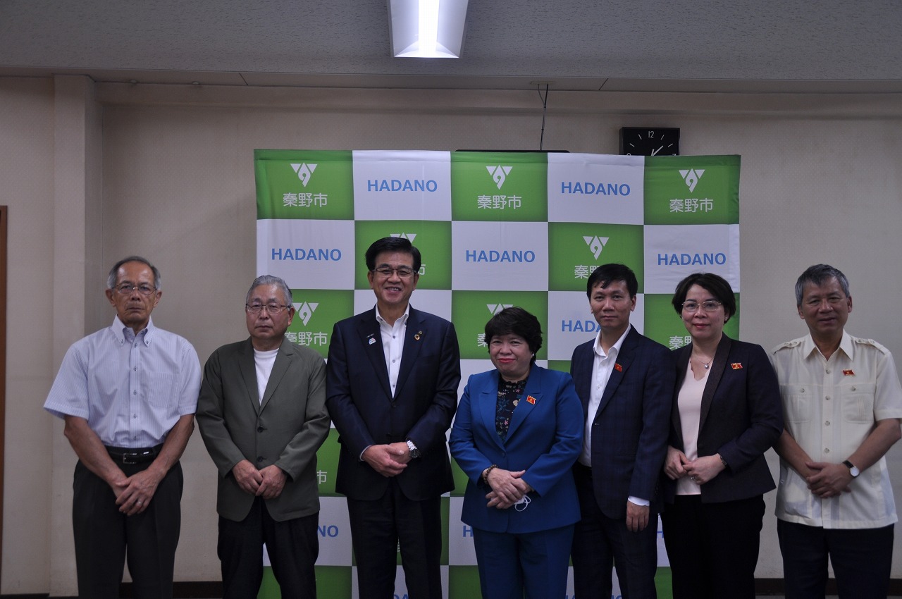 薄い緑と白の秦野市のチェック柄のバックパネルを背景に、スーツ姿の市長を中心とした7人の人々が横一列に並んでカメラに向かっている写真